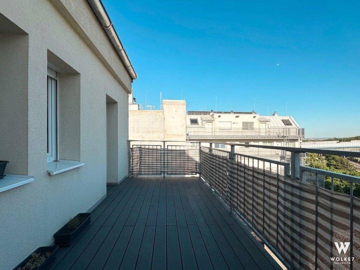 Langer Balkon mit Holzboden und weitem Blick über die städtische Umgebung unter blauem Himmel.