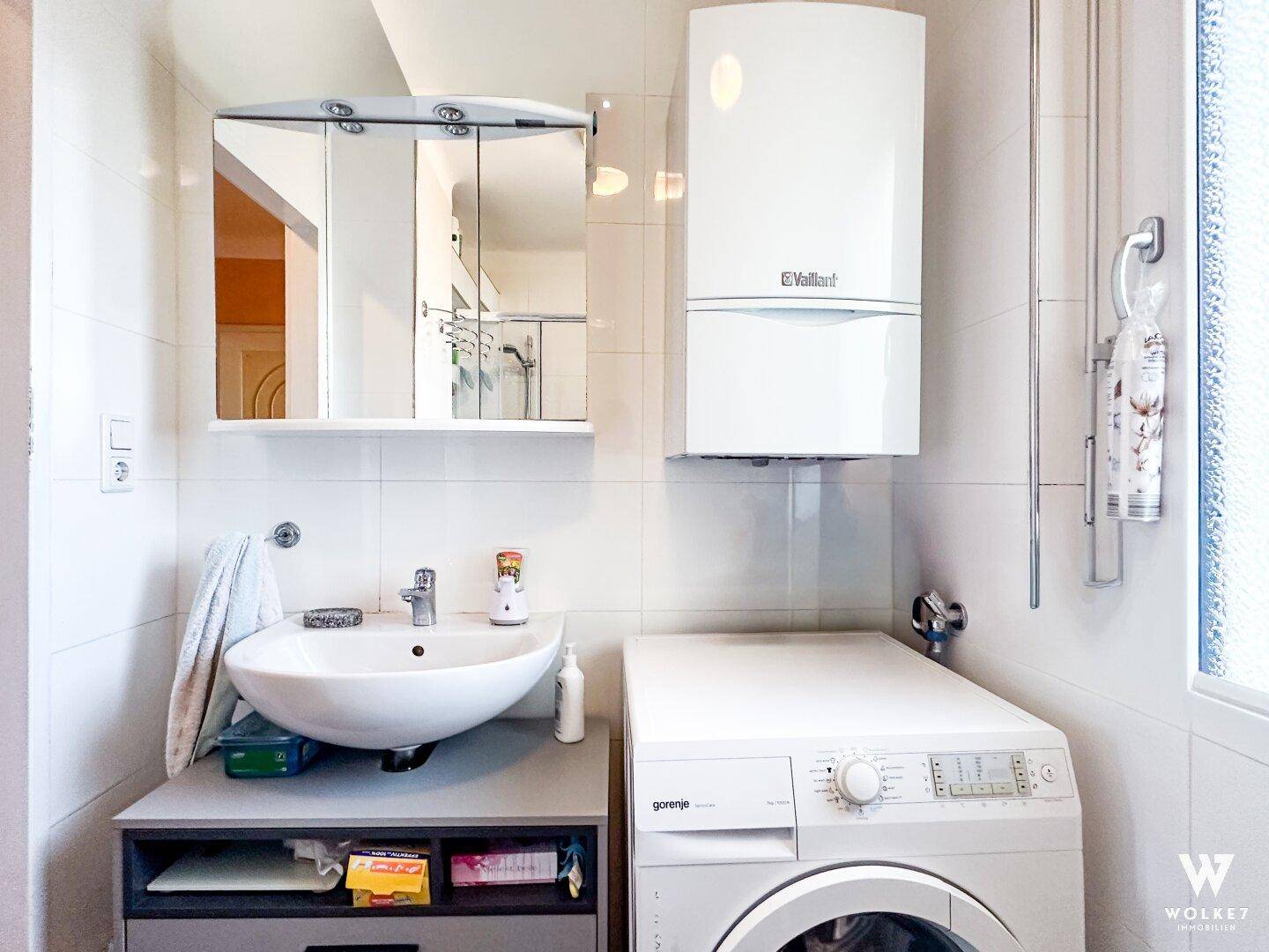 Badezimmer mit Waschbecken, Spiegelschrank, Waschmaschine und Warmwasserbereiter an der Wand.