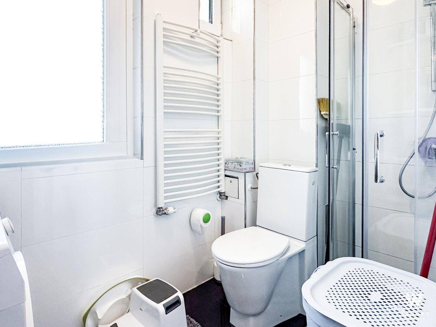 Helles Badezimmer mit WC, Duschkabine, Handtuchheizkörper und weißen Fliesen an den Wänden.
