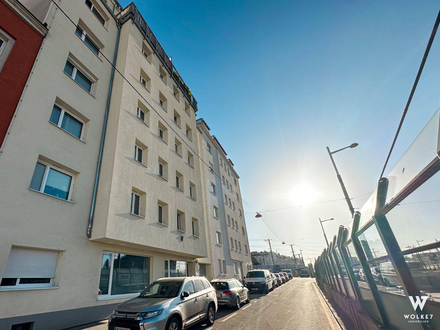 Mehrstöckiges Wohngebäude an einer Straße mit geparkten Autos unter strahlend blauem Himmel.
