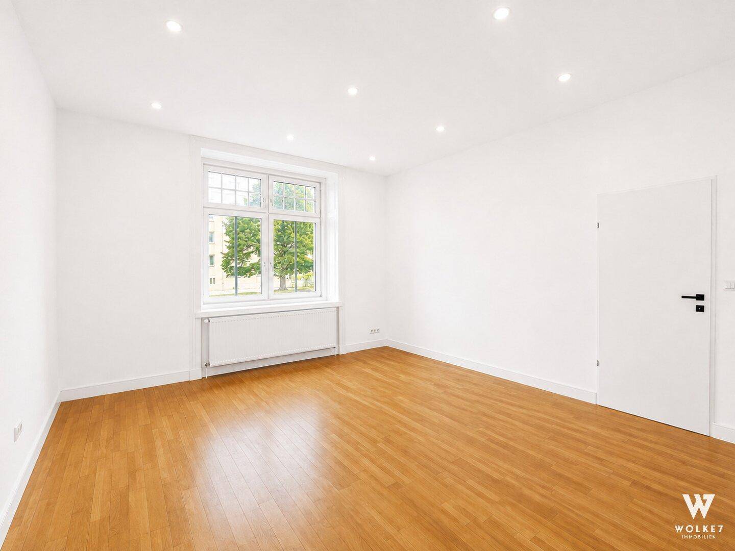 Geräumiges, leeres Zimmer mit Holzboden und großem Fenster, ideal für individuelle Gestaltung.