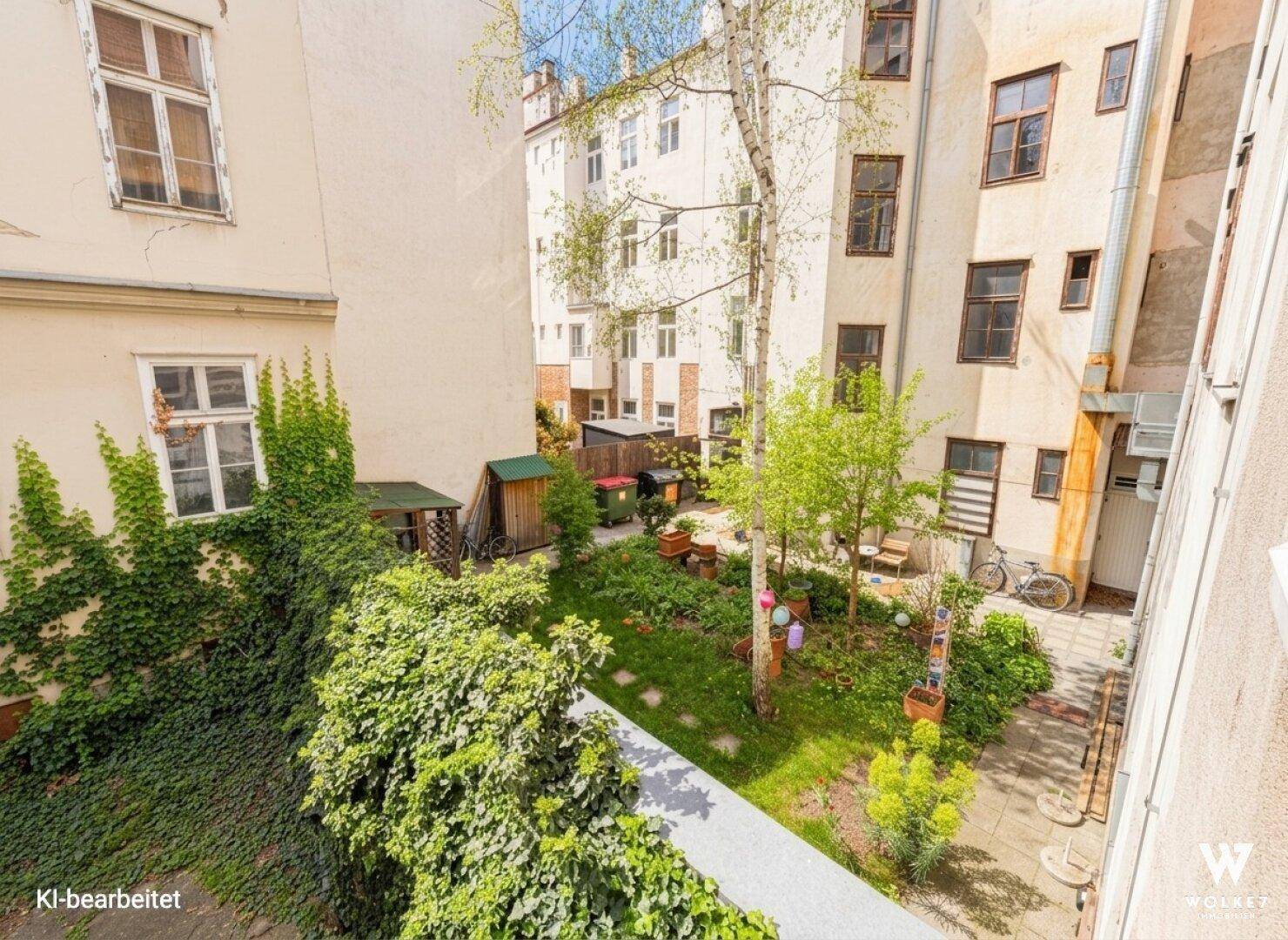 Blick auf einen grünen, liebevoll gestalteten Innenhofgarten mit Bäumen, Rasenflächen und Sitzgelegenheiten, umgeben von Altbauten.