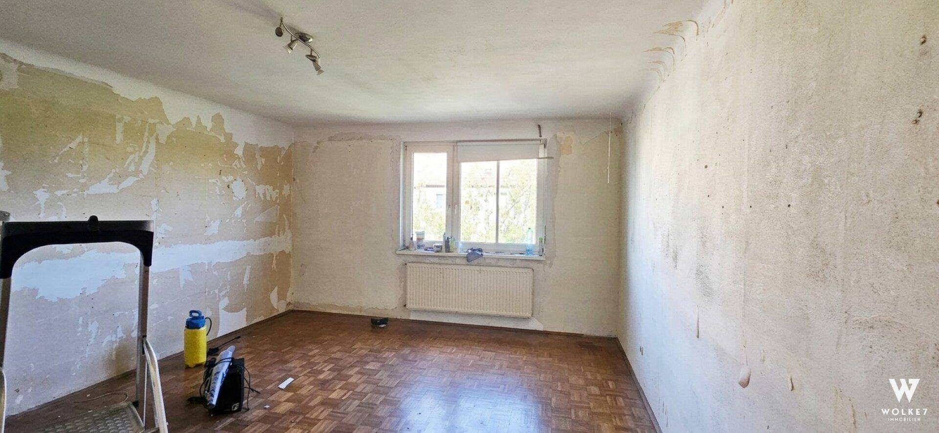 Leerer Raum im Renovierungszustand mit Fenster, Heizkörper und Parkettboden.