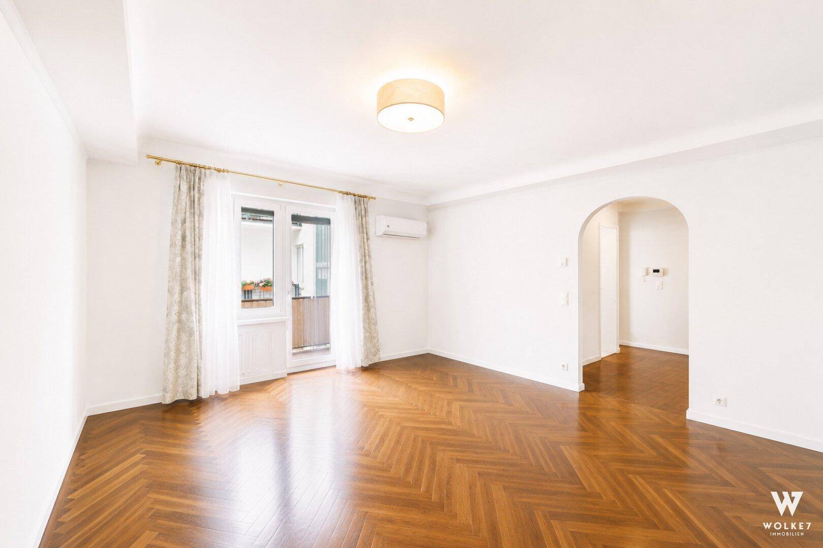 Geräumiges, leeres Wohnzimmer mit Fischgrätparkett, Balkonzugang und einem eleganten Bogen zu weiteren Räumen.