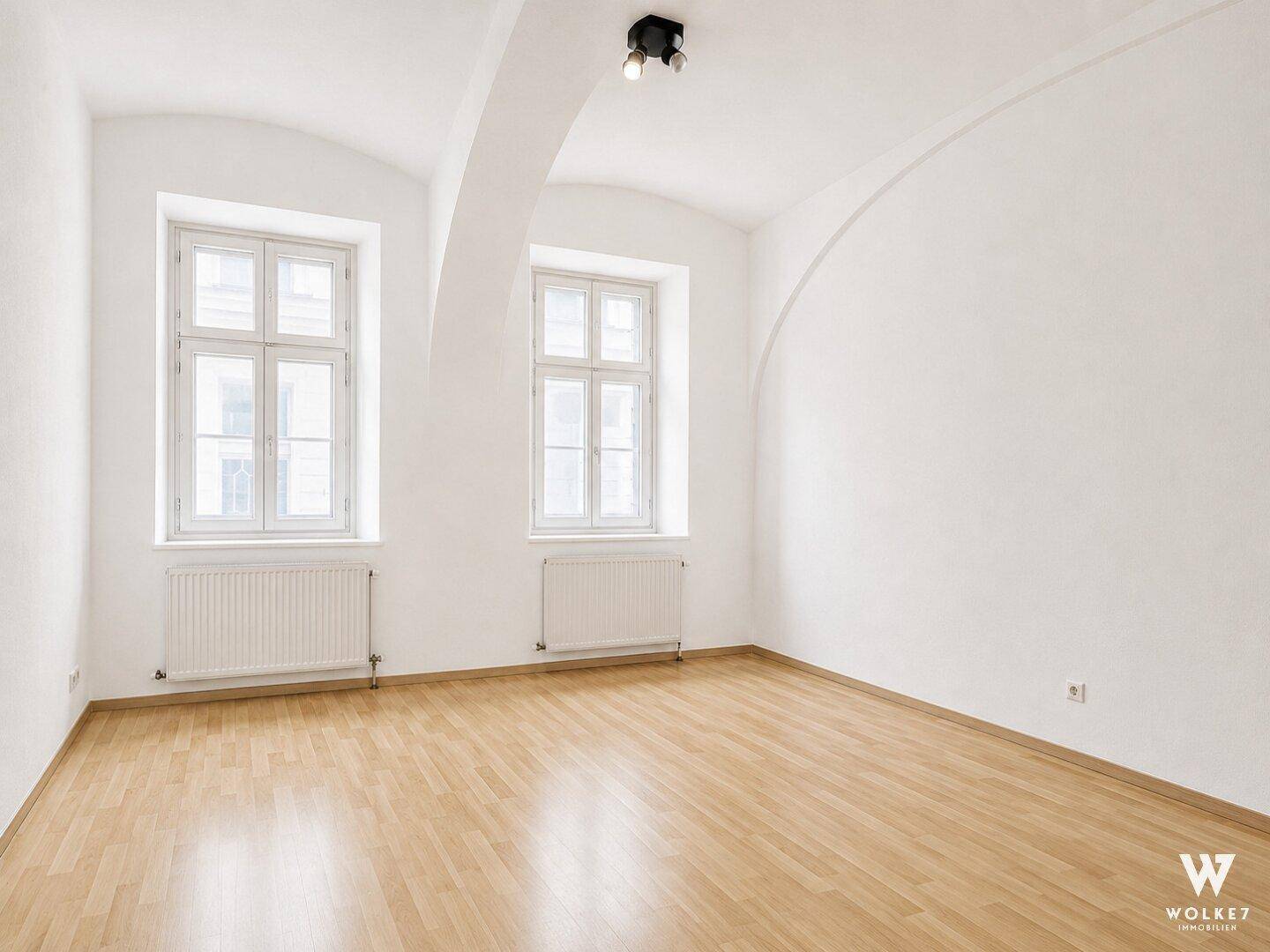 Leeres Wohnzimmer mit hellen Wänden, Holzboden und hohen Fenstern, bereit zur individuellen Gestaltung.
