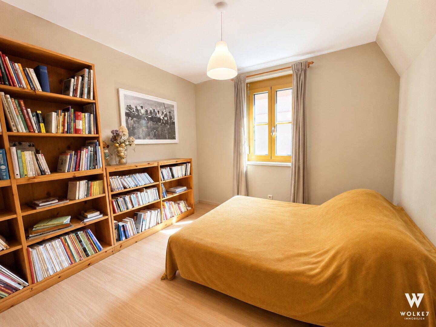 Helles Schlafzimmer mit Bücherregalen, Holzboden und Fenster mit Blick ins Grüne und Dachschräge.