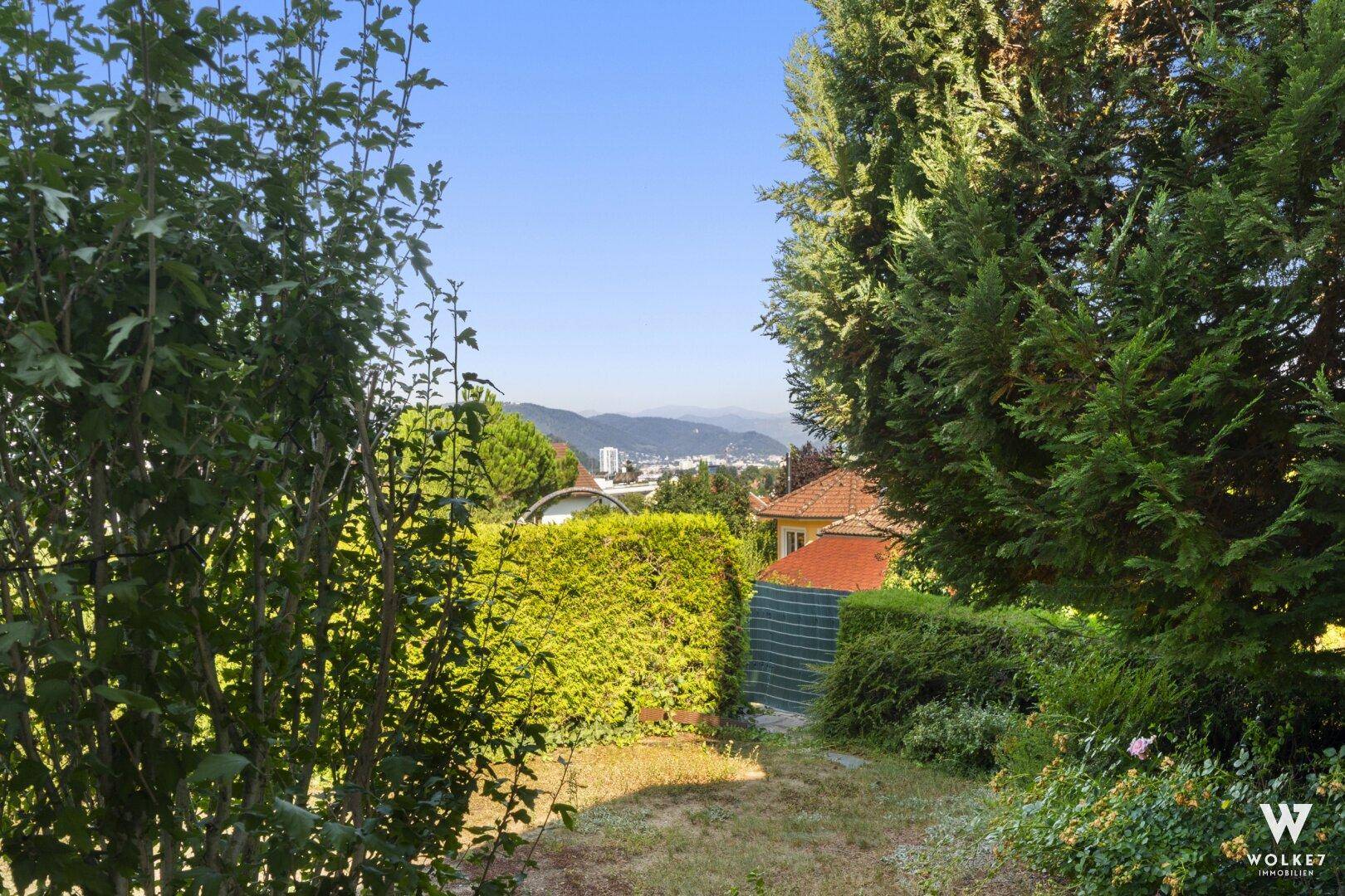 Gartenbereich mit Hecken, Bäumen und Fernblick auf die Stadt und Berge.