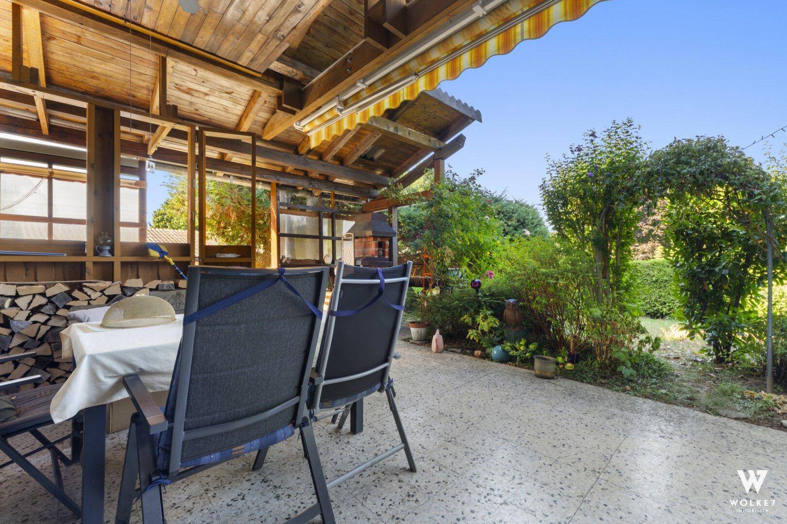 Überdachte Terrasse mit Gartenmöbeln, Holzstapel und Blick ins Grüne.