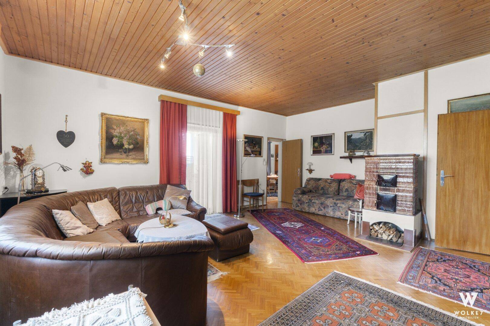 Großes Wohnzimmer mit Holzbalkendecke, Kachelofen und Zugang zum Balkon.