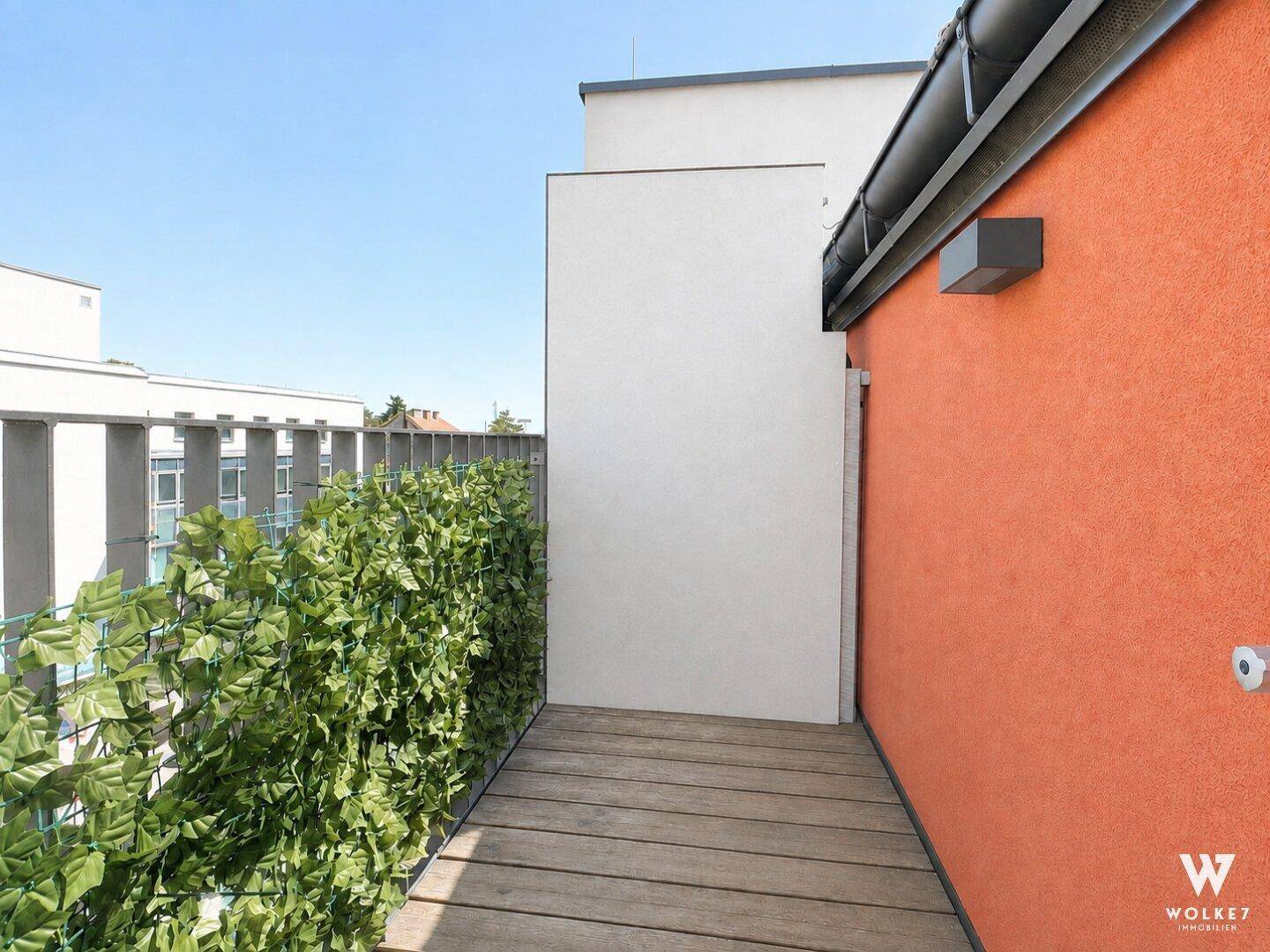 Schmaler Balkon mit Holzboden und grüner Sichtschutzhecke unter blauem Himmel.