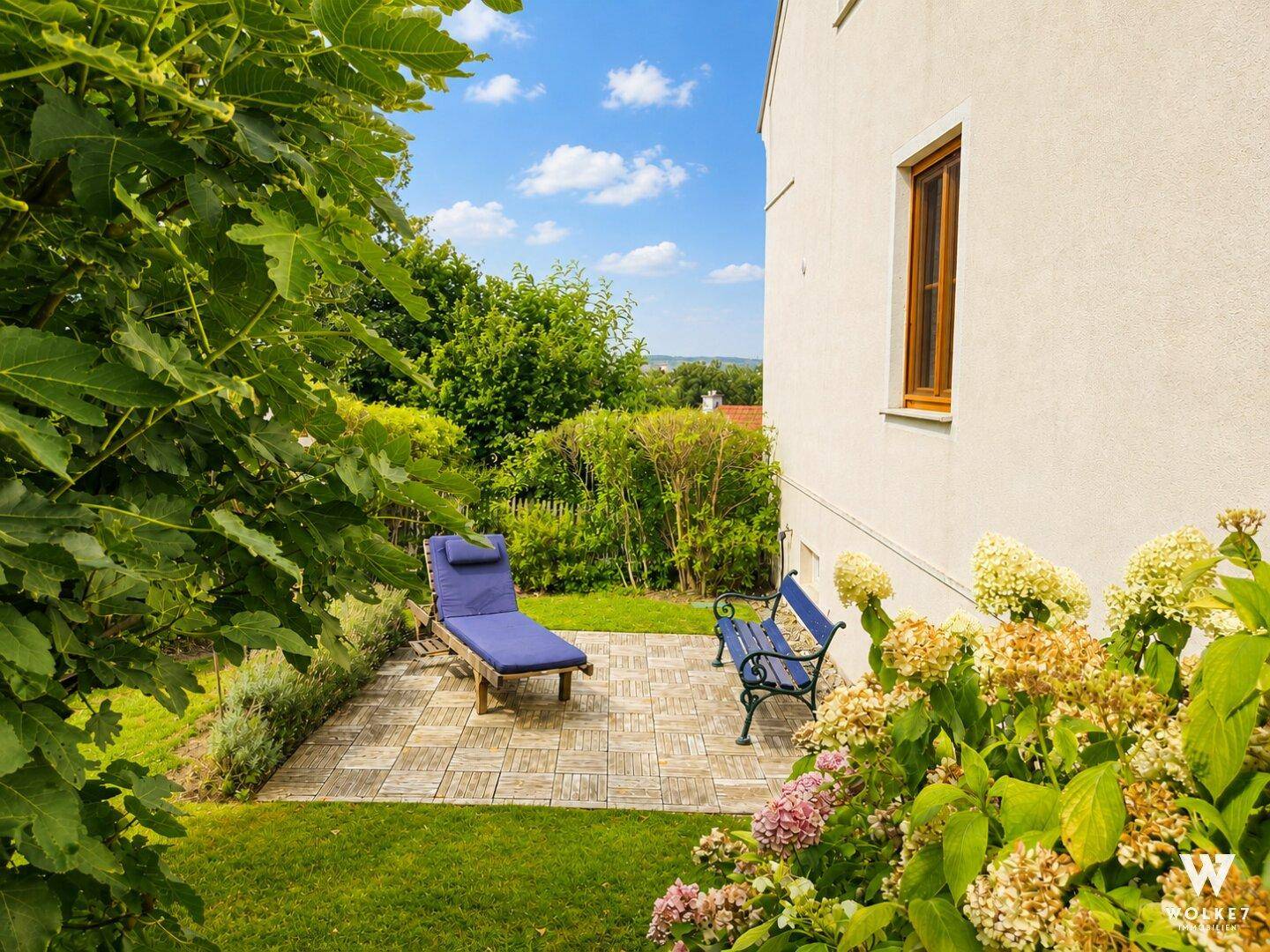 Ruhige gepflasterte Terrasse mit Liegestuhl und Sitzbank, umgeben von üppigem Grün und Blumen.
