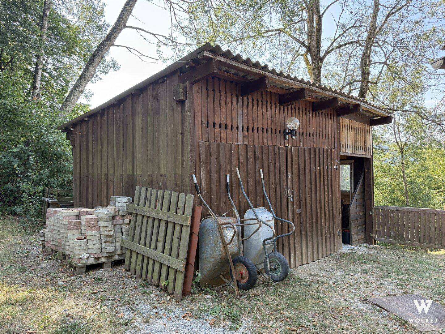 Hölzerner Geräteschuppen mit Schubkarren und Baumaterialien im Gartenbereich.