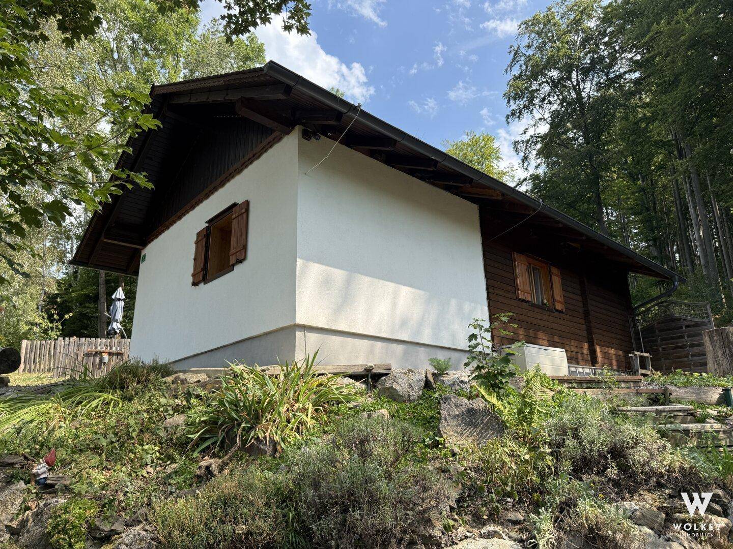 Seitenansicht des Hauses mit weißer Fassade und Holzverkleidung, umgeben von Pflanzen.