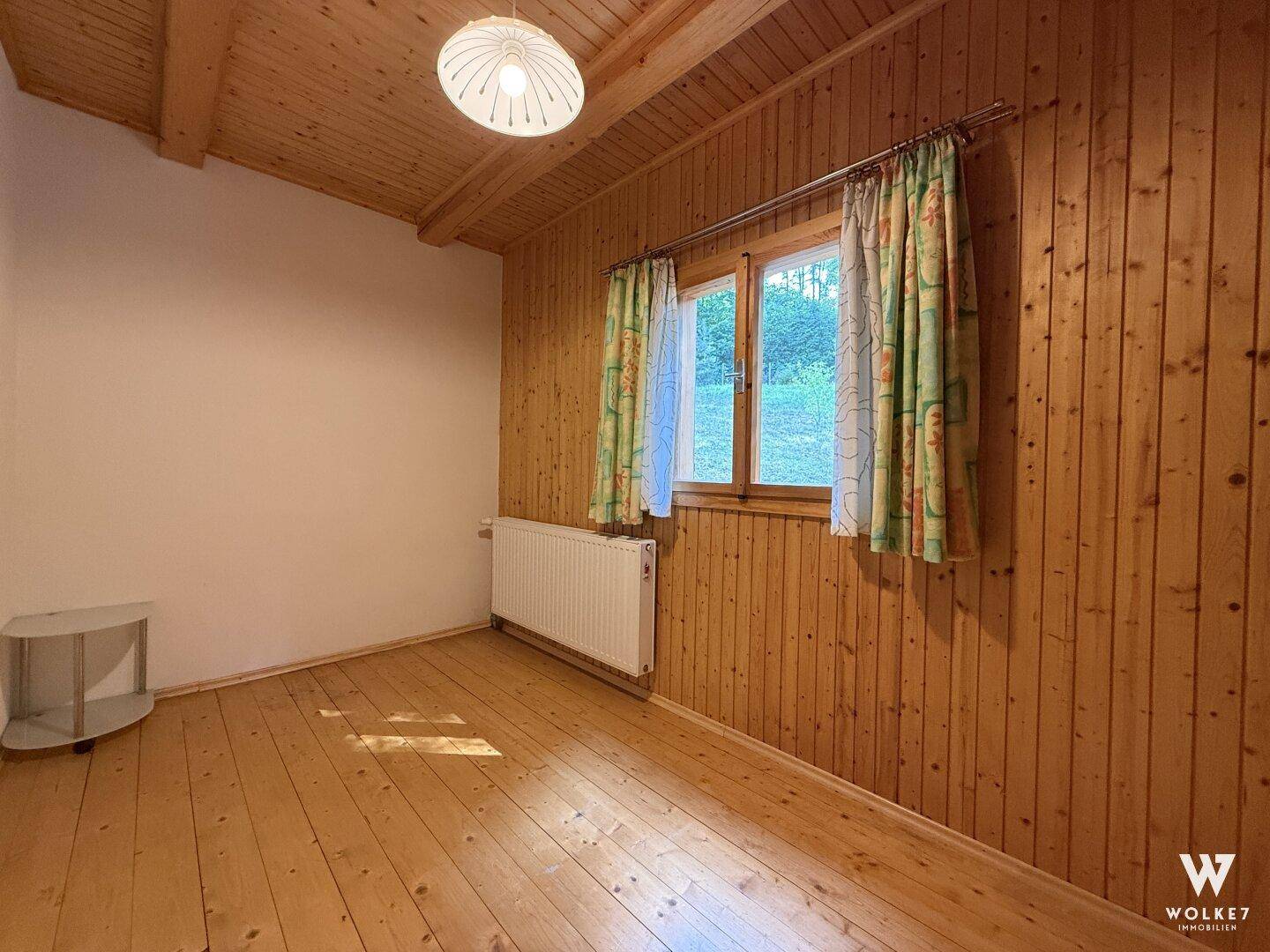 Heller Wohnraum mit Holzwänden, Holzboden und einem Fenster mit Blick ins Grüne.