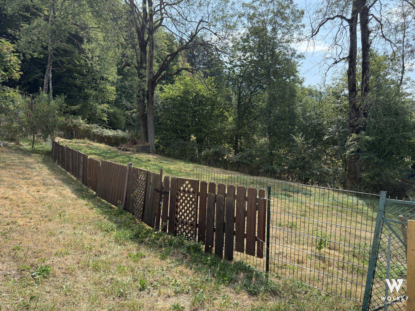 Eingezäunter Gartenbereich mit Rasenfläche und Bäumen im Hintergrund.