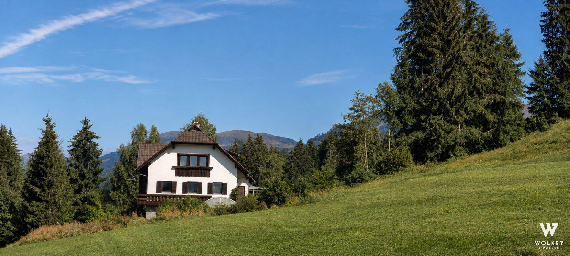 Charmantes Haus mit weißer Fassade und Satteldach, eingebettet in eine grüne Hügellandschaft mit hohen Bäumen.