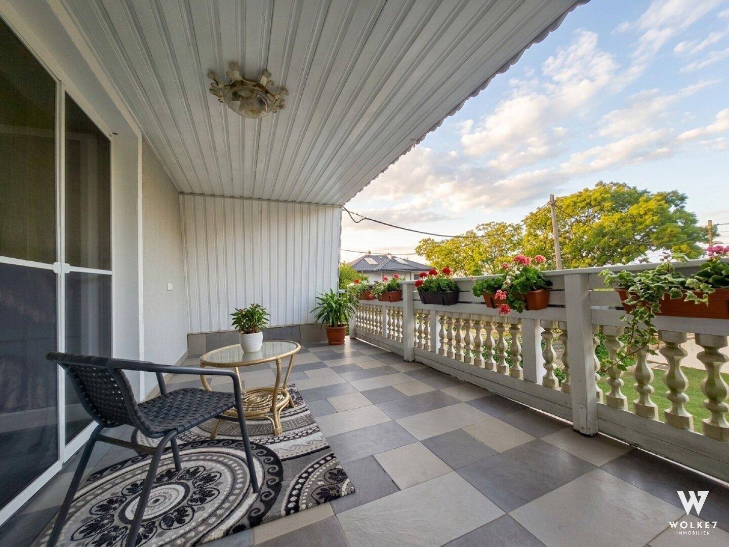 Überdachter Balkon mit Fliesenboden, Sitzgelegenheiten und Blumenkästen.