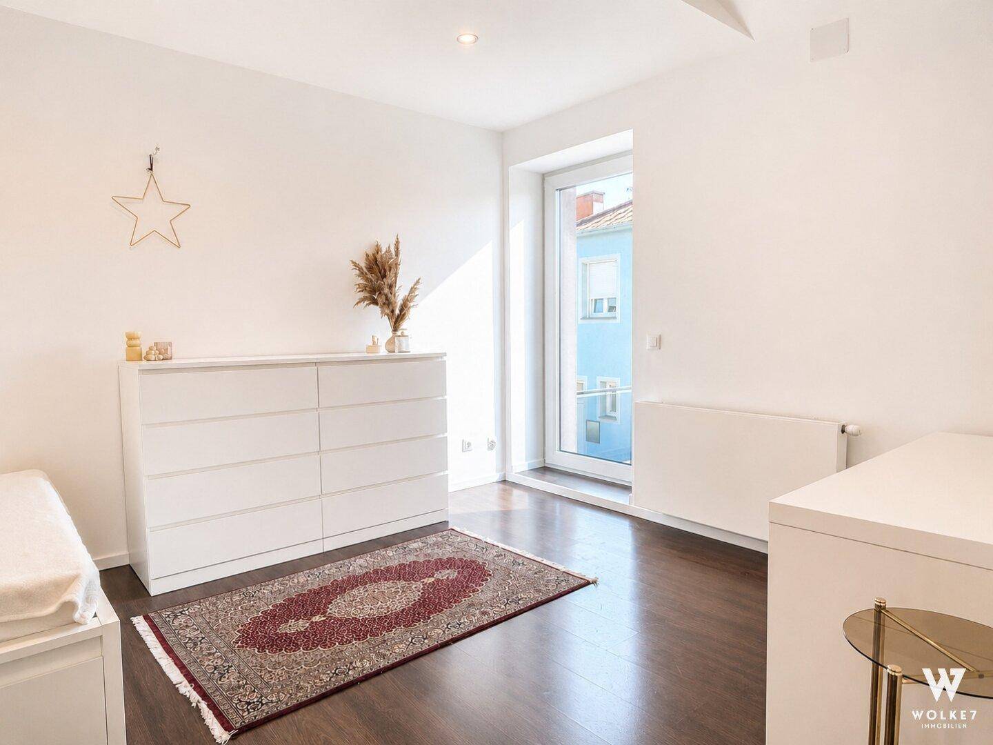 Helles Schlafzimmer mit dunklem Holzboden, weißer Kommode und Zugang zum Balkon.