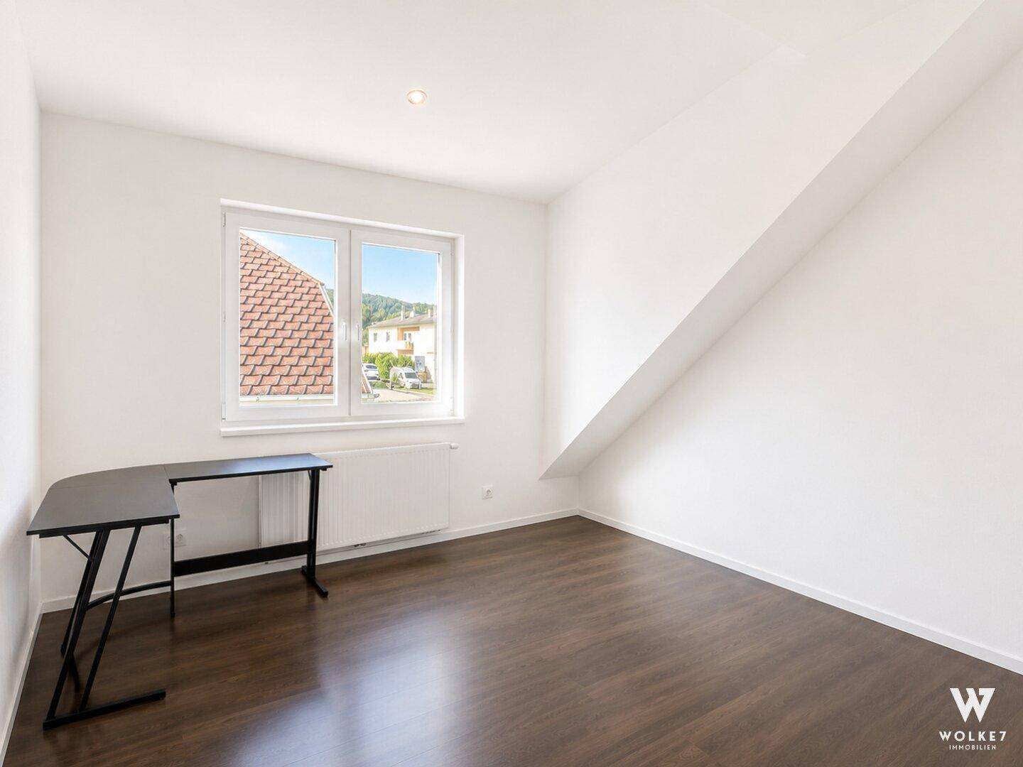 Helles Schlafzimmer oder Büro mit Dachschräge, dunklem Boden und Fensterblick.