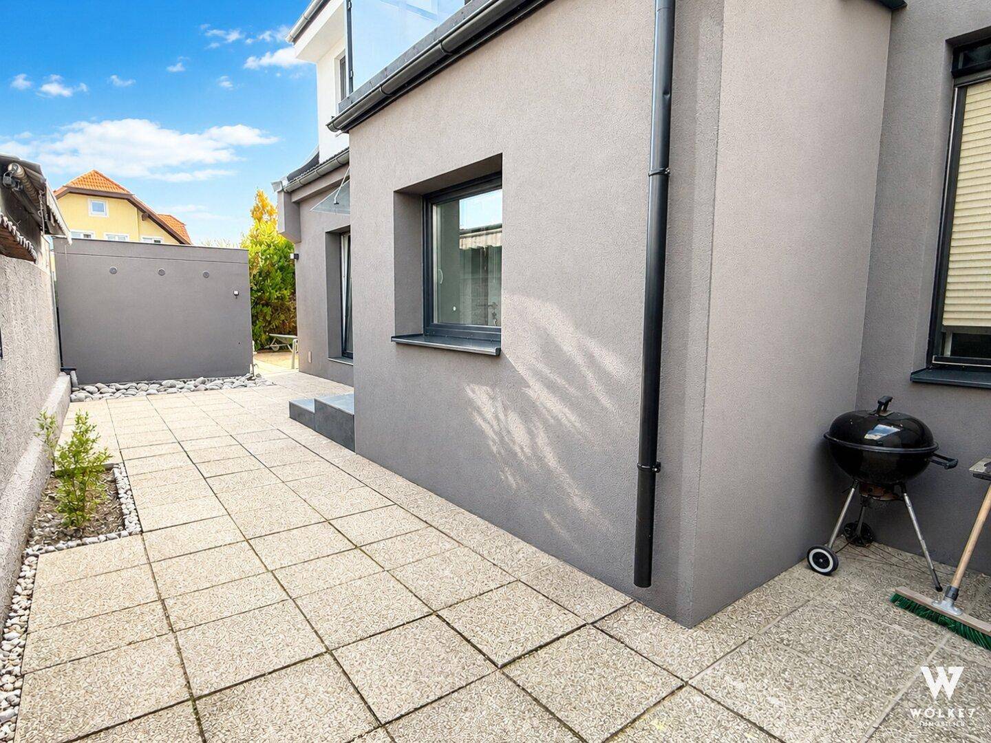Gepflasterter Außenbereich mit Grill und kleiner Bepflanzung neben dem Haus.