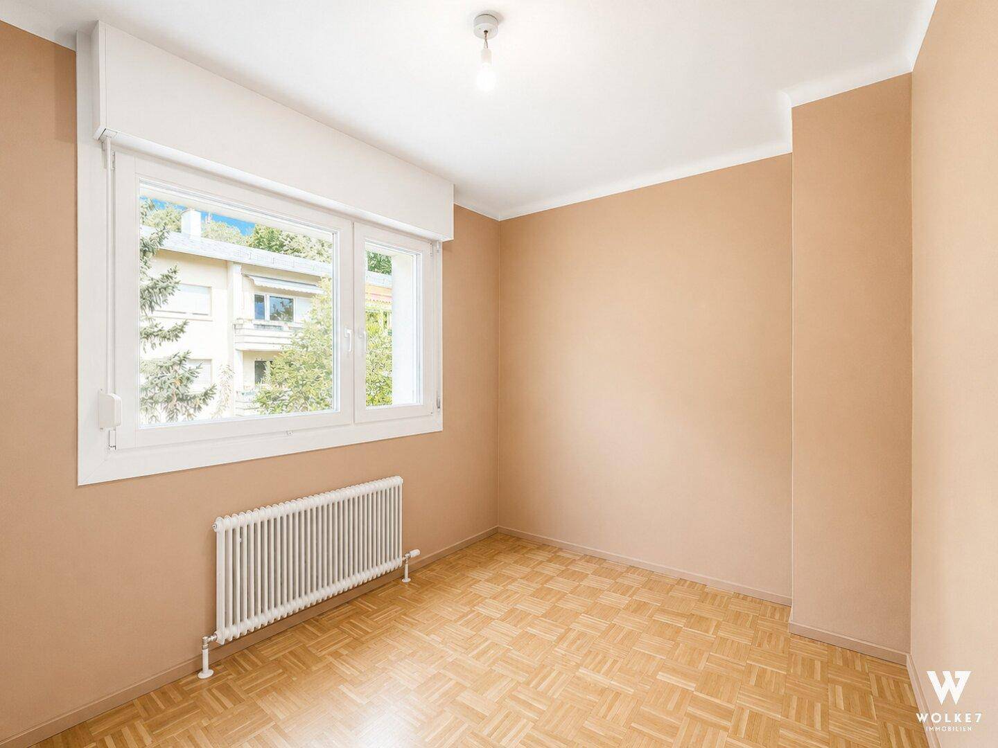 Leeres Schlafzimmer mit Parkettboden, beigen Wänden und einem großen Fenster für viel Tageslicht.