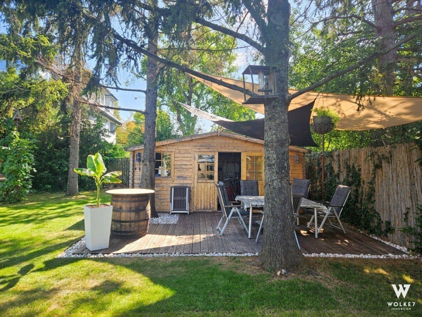 Gemütlicher Sitzbereich im Garten mit Holzterrasse, Gartenhaus und schattenspendenden Bäumen.