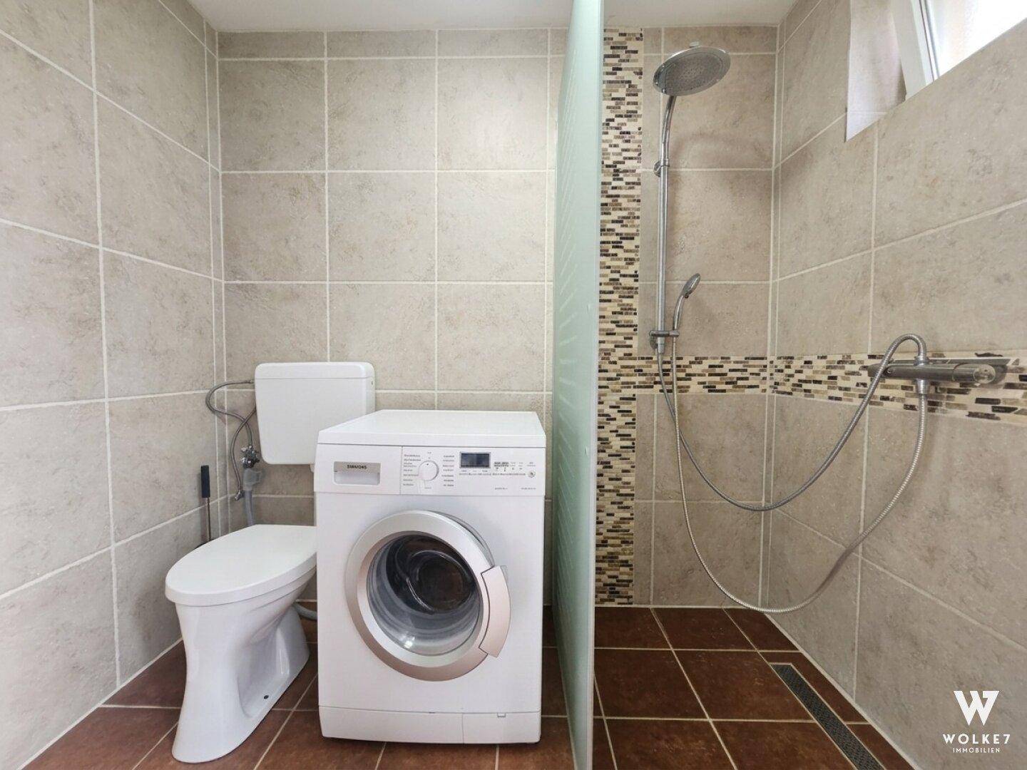 Funktionales Badezimmer mit Duschbereich, WC und Waschmaschine, hell gefliest.