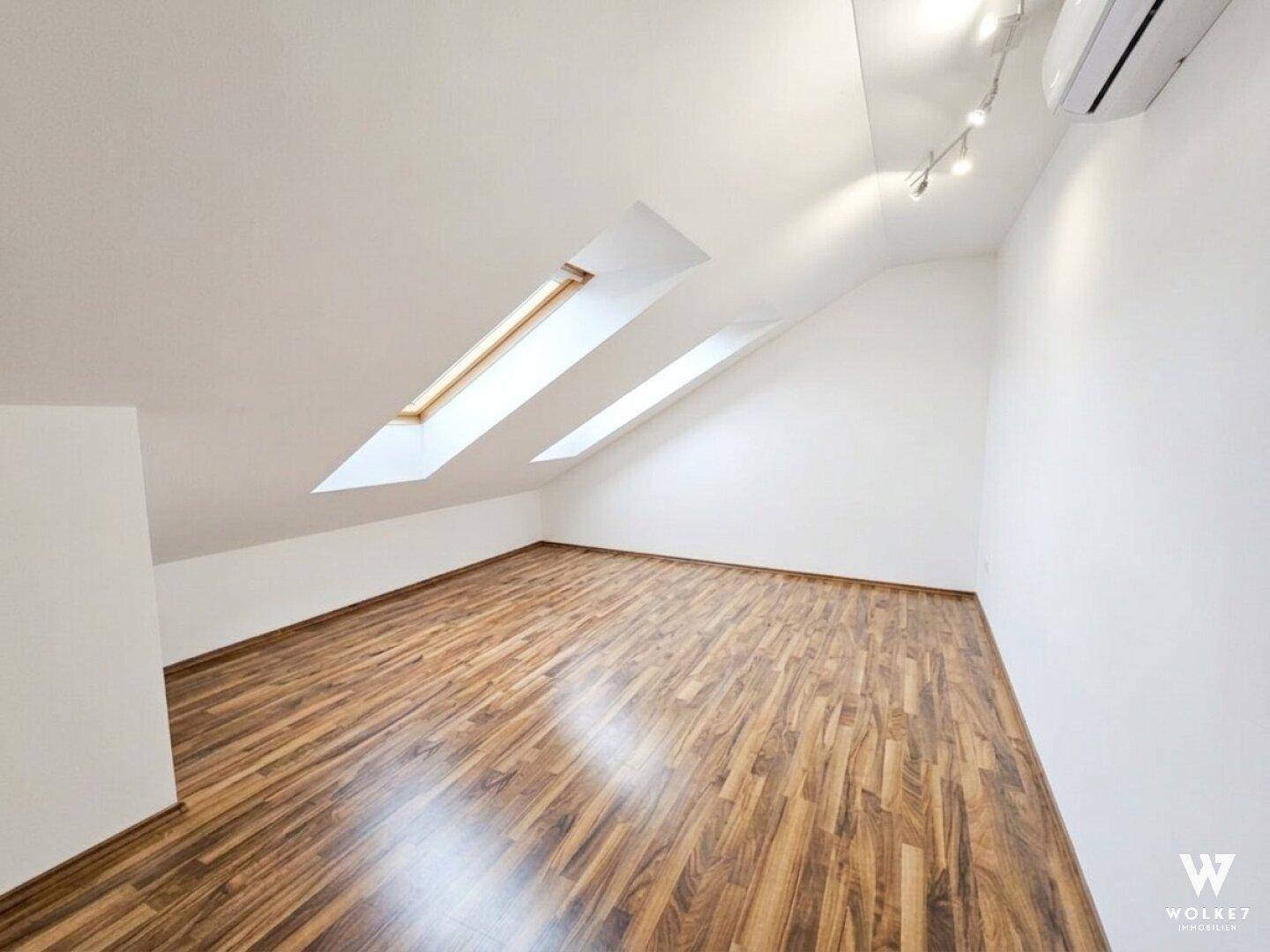 Helles Dachzimmer mit schrägen Wänden, Dachfenstern und warmem Laminatboden.