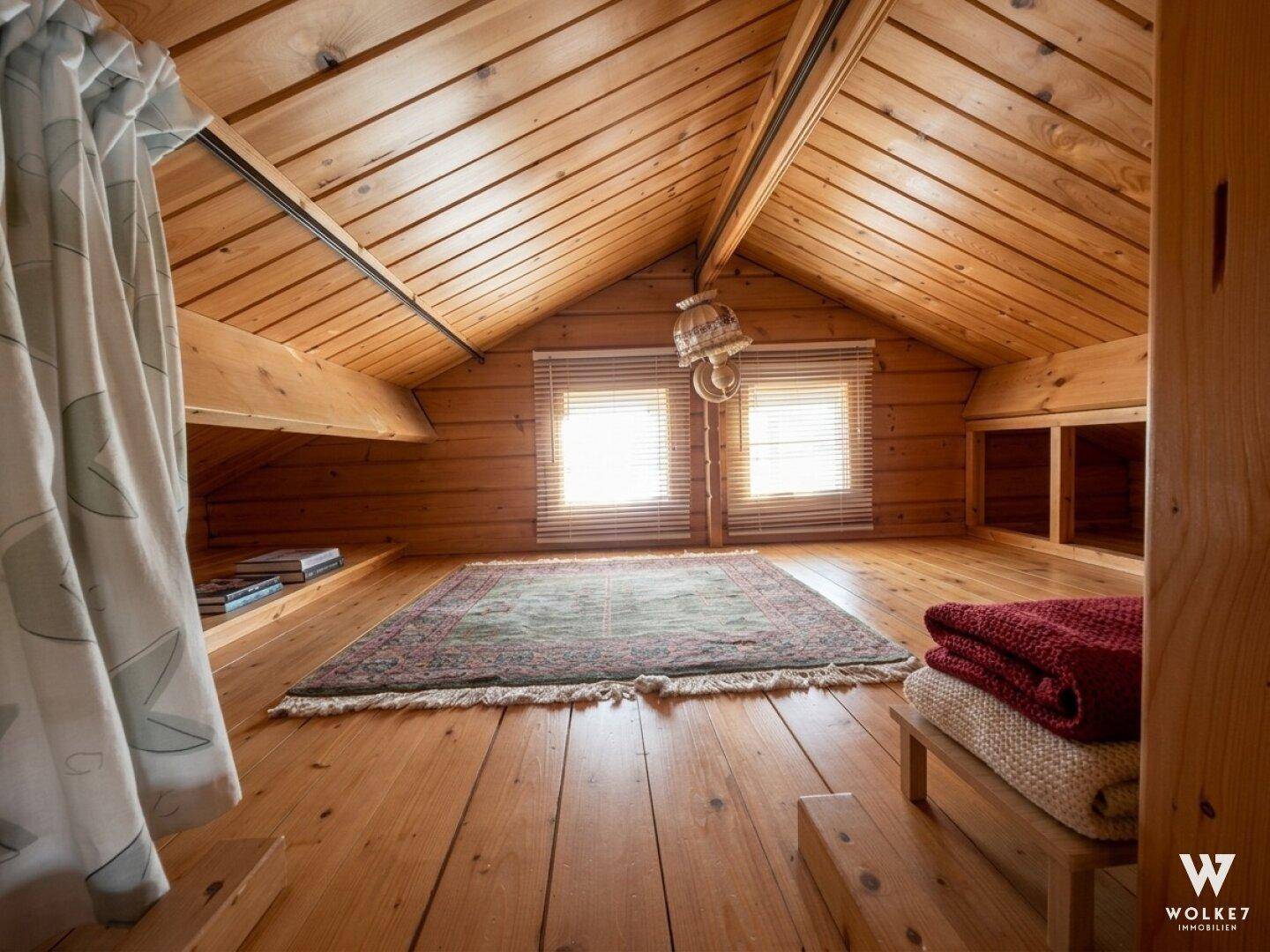 Heller Dachbodenbereich in der Holzhütte mit schrägen Holzdecken, Teppich und zwei kleinen Fenstern.