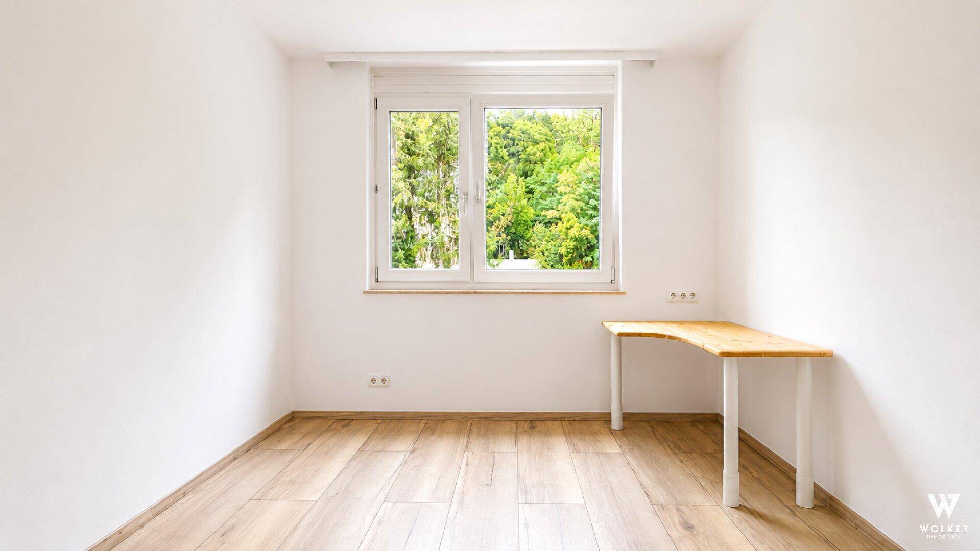 Freundliches Arbeitszimmer mit hellem Holzschreibtisch, Parkettboden und Blick ins Grüne.