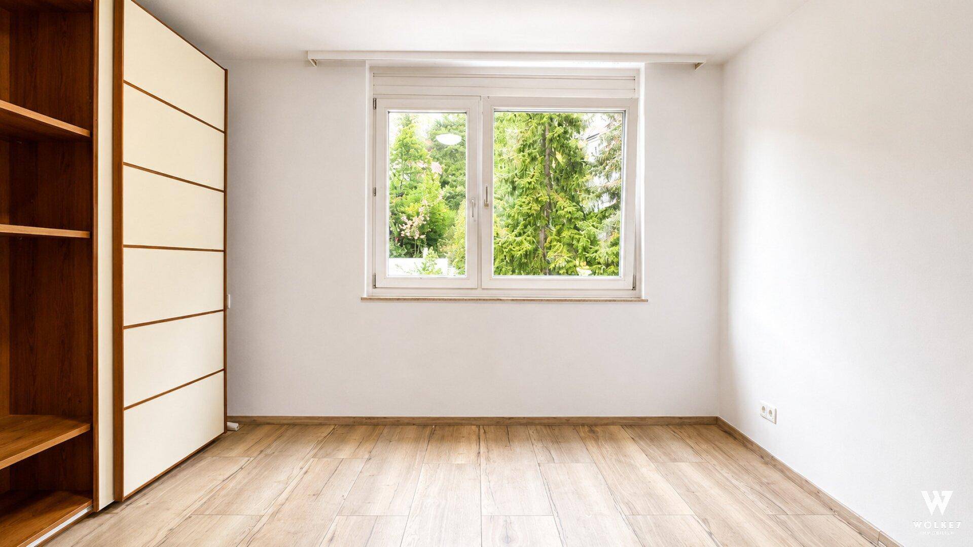 Helles Zimmer mit Holzfußboden, großem Fenster und integriertem Schrank, bietet Blick ins Grüne.