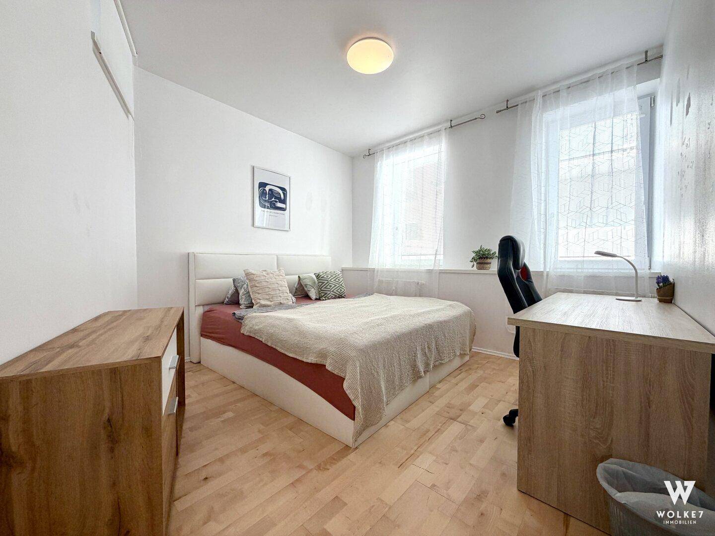 Schlafzimmer mit Doppelbett, Schreibtisch und hellem Holzboden, gut beleuchtet durch Fenster.