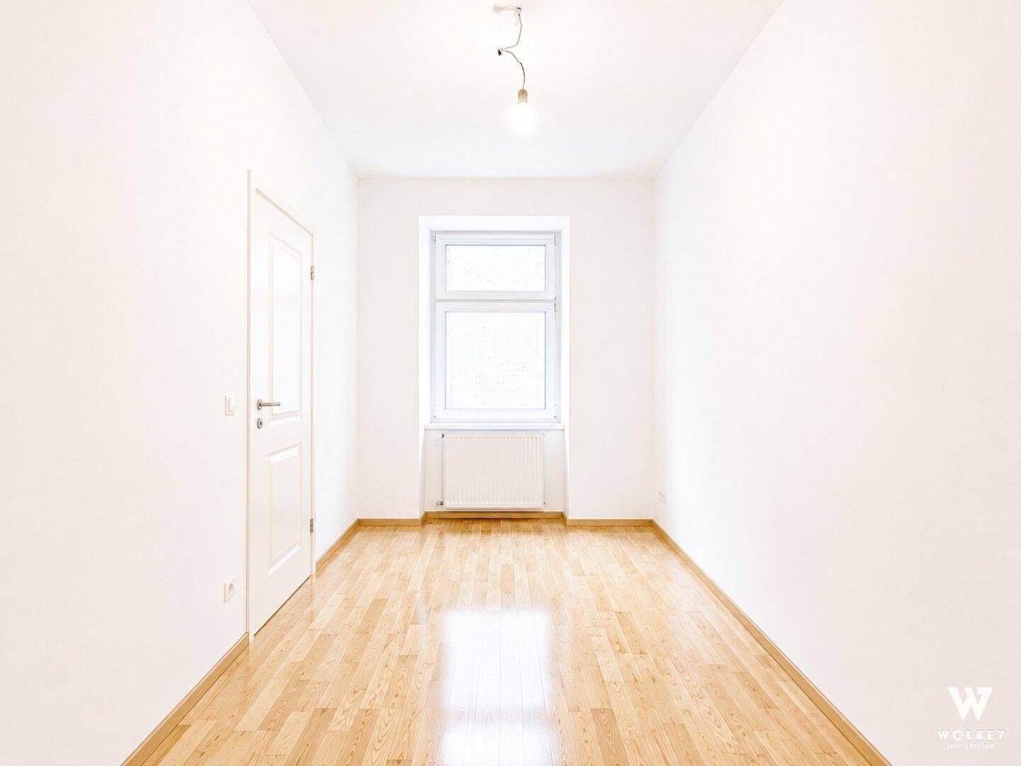 Leeres Zimmer mit hellem Parkettboden und einem großen Fenster für viel Tageslicht.