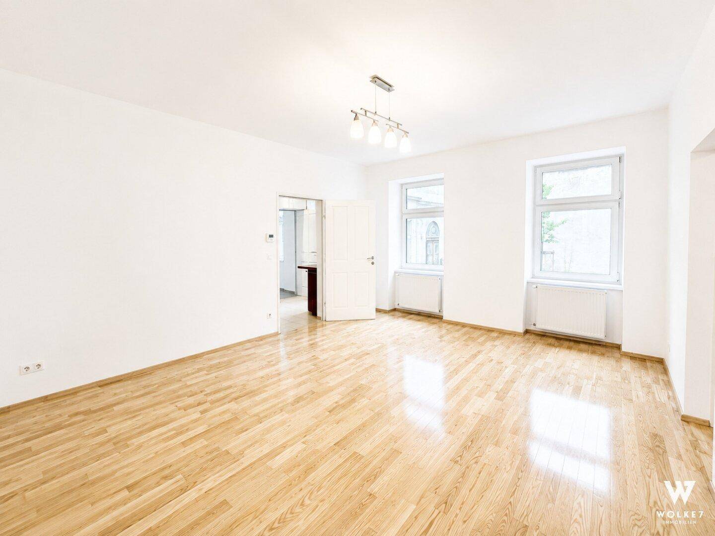 Leeres, helles Wohnzimmer mit glänzendem Parkettboden und großen Fenstern.