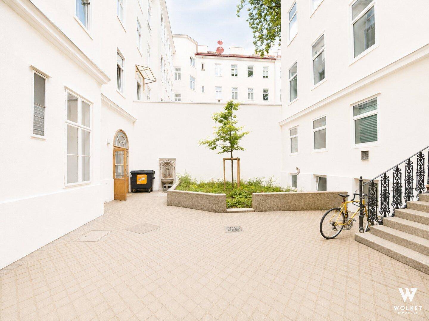 Gepflasterter Innenhof mit jungem Baum, Sitzgelegenheit und Zugang zum Gebäude.