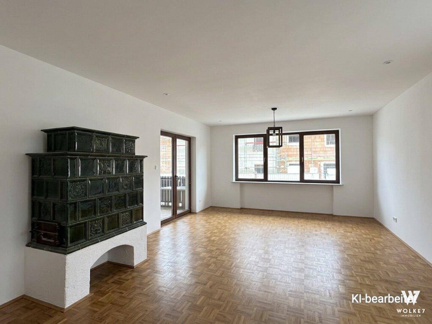 Geräumiges Wohnzimmer mit Fischgrätparkett, großen Fenstern und einem traditionellen Kachelofen.