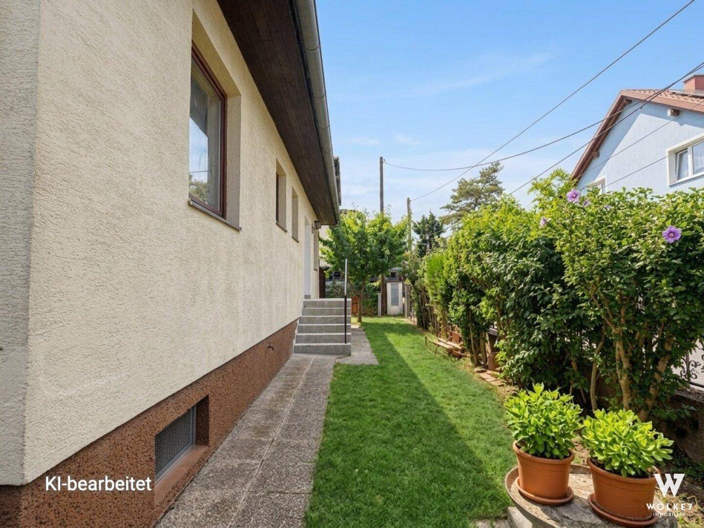Schmaler Gartenweg entlang der Hausseite mit grüner Hecke und Topfpflanzen.