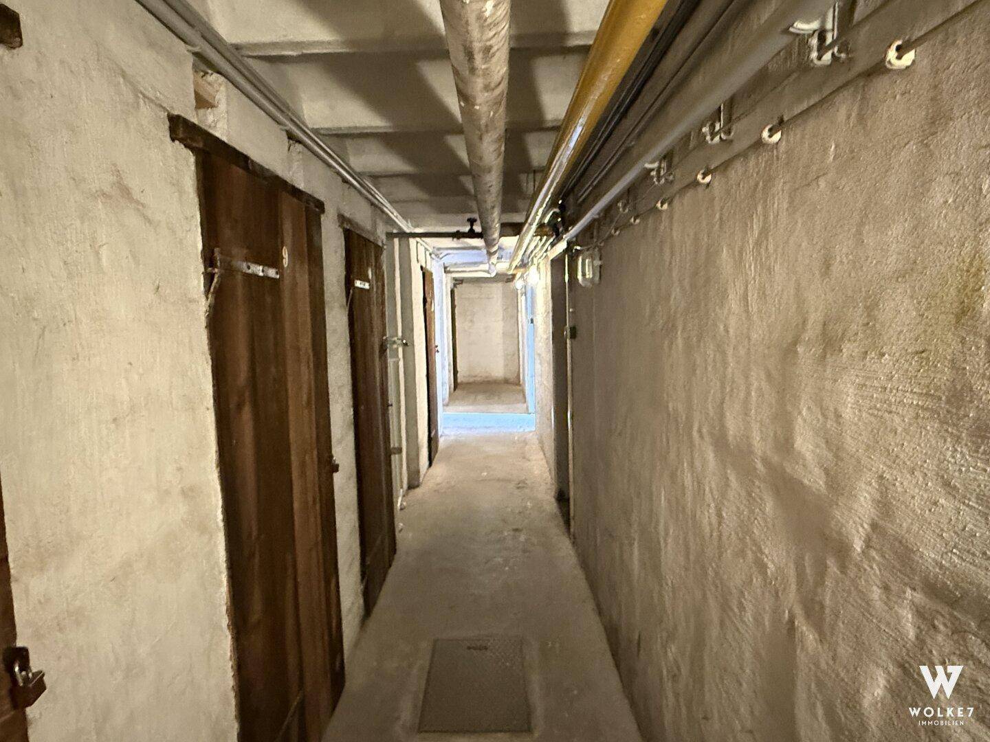 Langer Kellerflur mit einzelnen Abteilen und sichtbaren Rohren an der Decke.