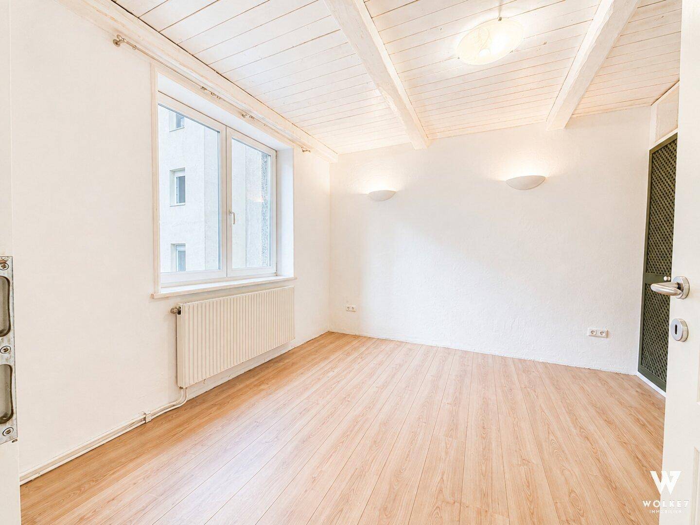 Helles Zimmer mit Holzbalkendecke, Holzboden und Fensterblick nach draußen.