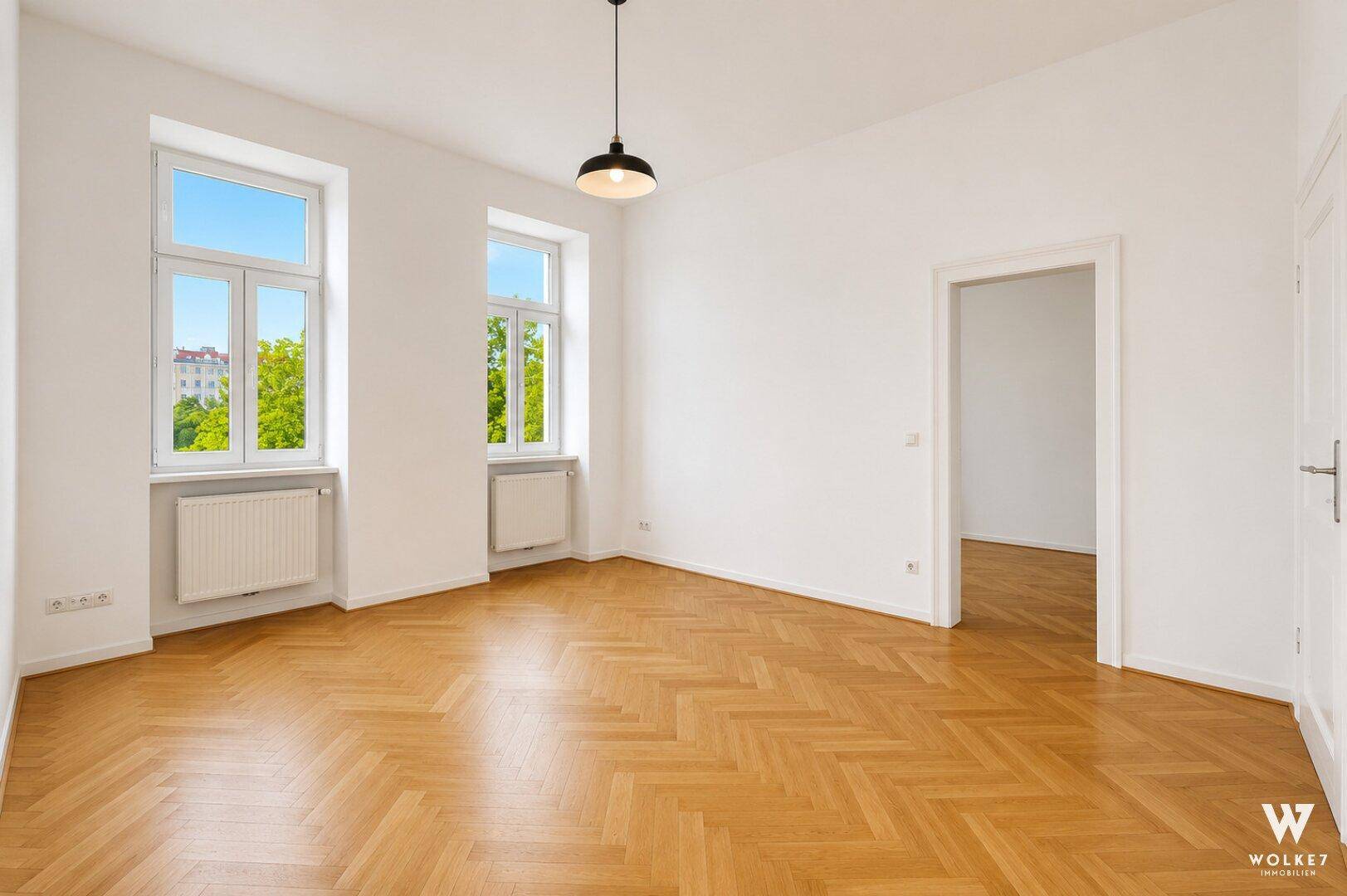 Heller Wohnraum mit Fischgrätparkett, hohen Decken und zwei Fenstern mit Blick ins Grüne.