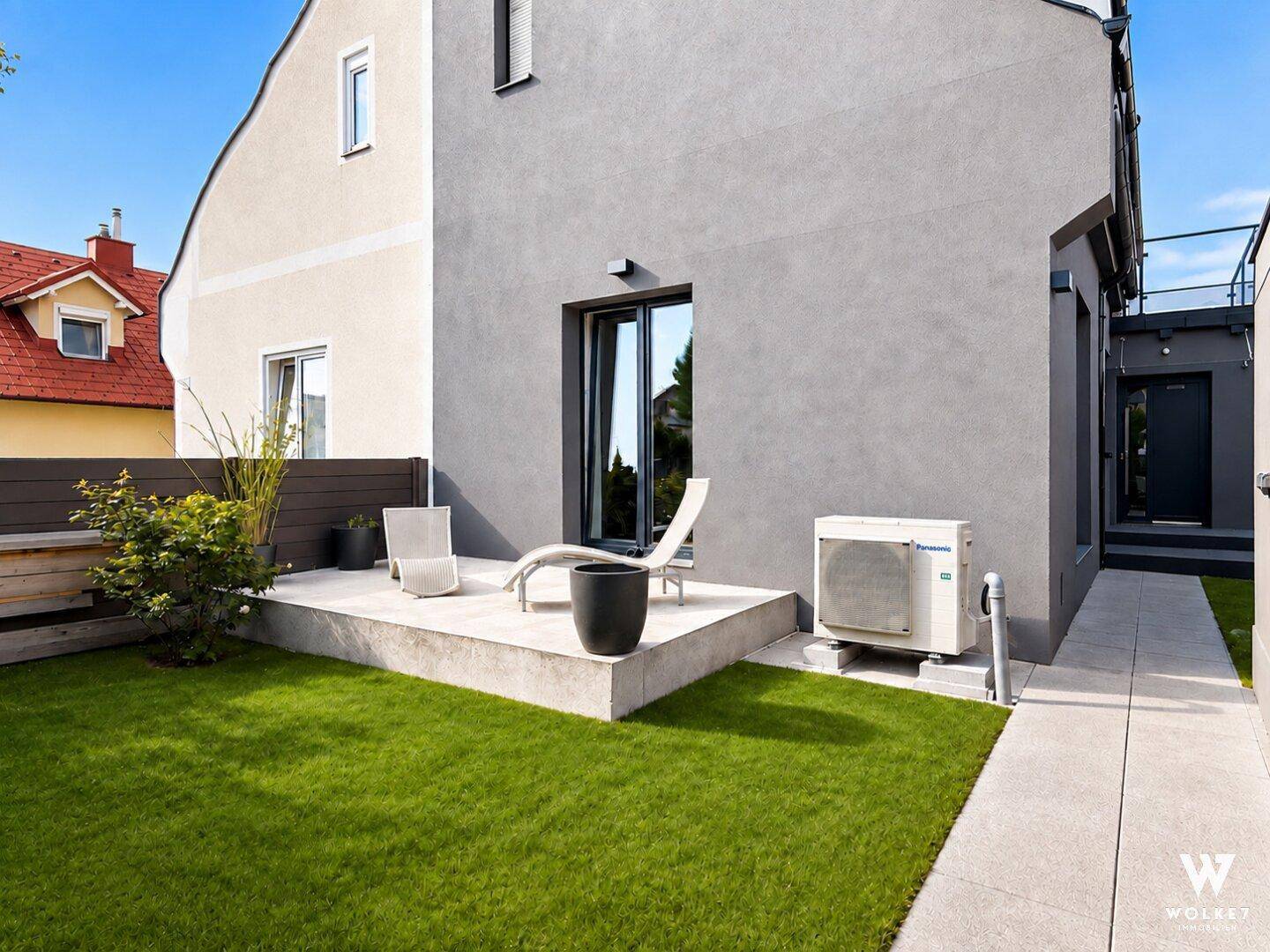 Moderner Gartenbereich mit kleiner Rasenfläche, Beton-Terrasse und Lounge-Möbeln neben dem Haus.