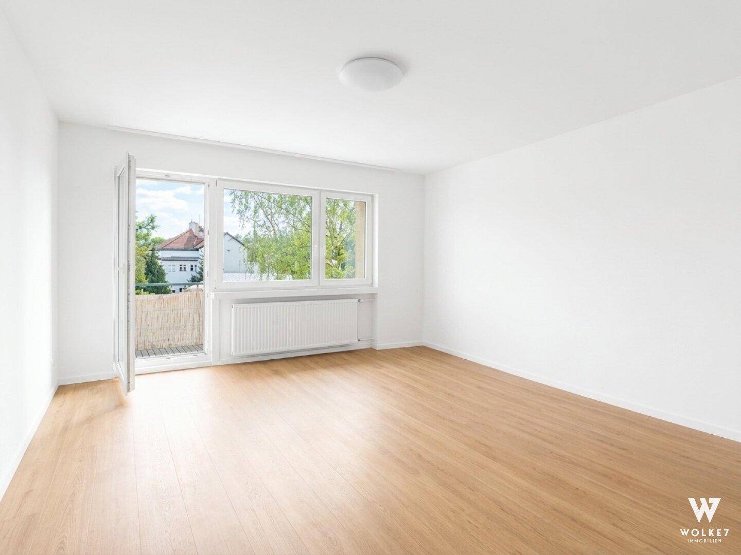 Geräumiges, unmöbliertes Zimmer mit hellem Holzboden und Fensterfront zum Balkon.