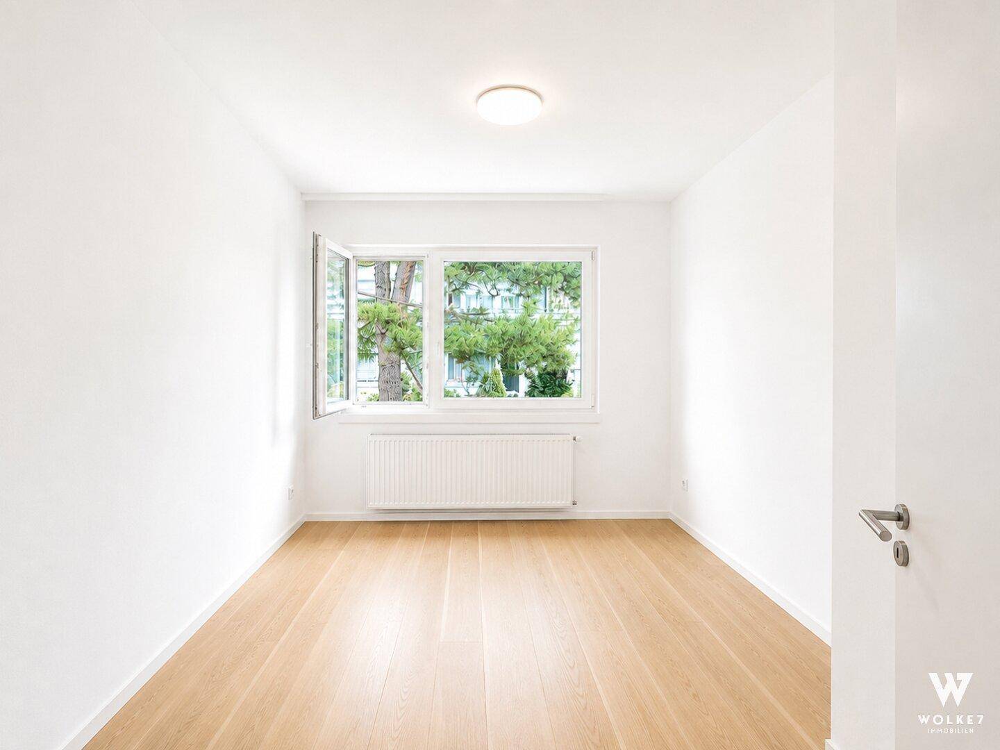 Helles, unmöbliertes Zimmer mit Holzboden und Fenster mit Blick ins Grüne.