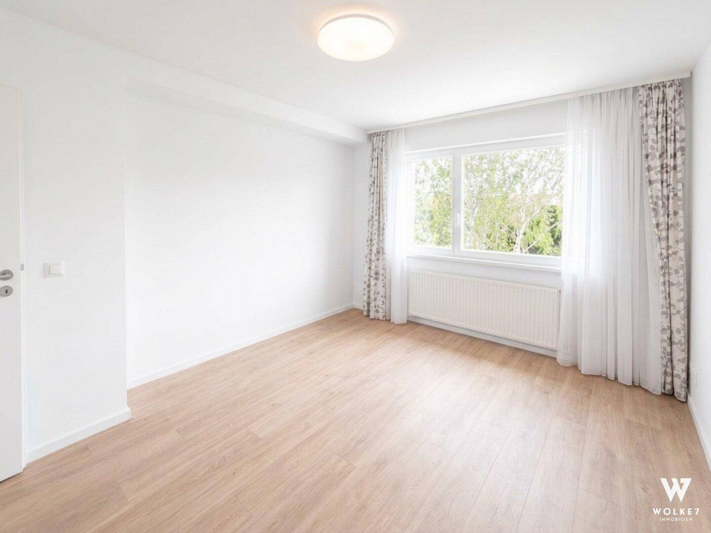 Geräumiges, leeres Zimmer mit hellem Holzboden und großen Fenstern.