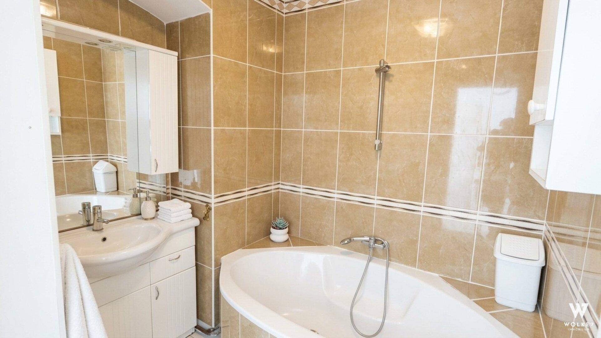 Gefliestes Badezimmer mit beigen Fliesen, Eckbadewanne und Waschbecken mit Spiegelschrank.