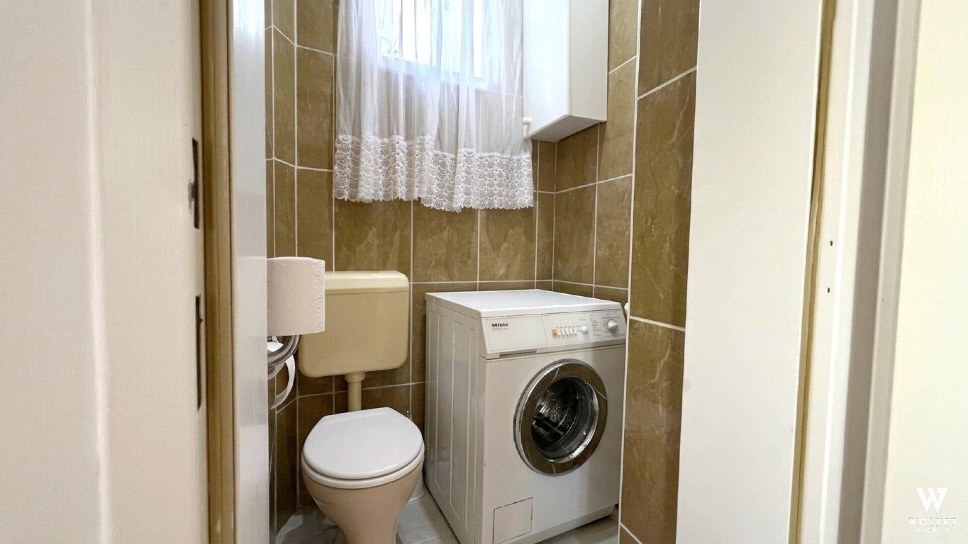 Kleiner WC-Raum mit Toilette, Waschmaschine und Fenster, komplett gefliest.