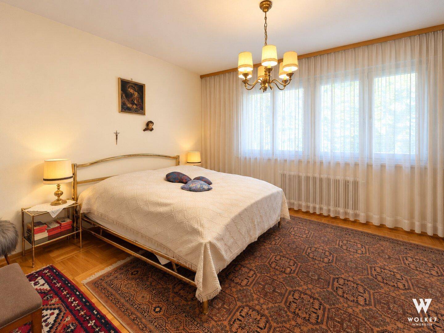 Traditionelles Schlafzimmer mit Doppelbett, Nachttischen und großen Fenstern mit Vorhängen.