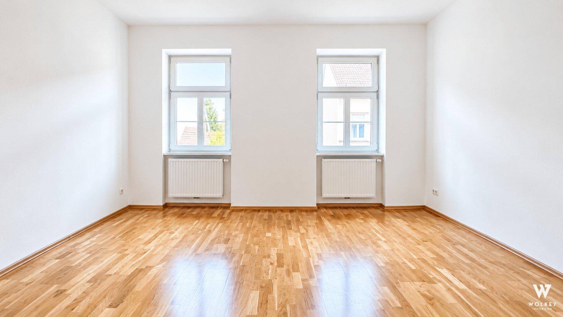 Geräumiges, helles Zimmer mit klassischem Parkettboden und zwei großen Fenstern.