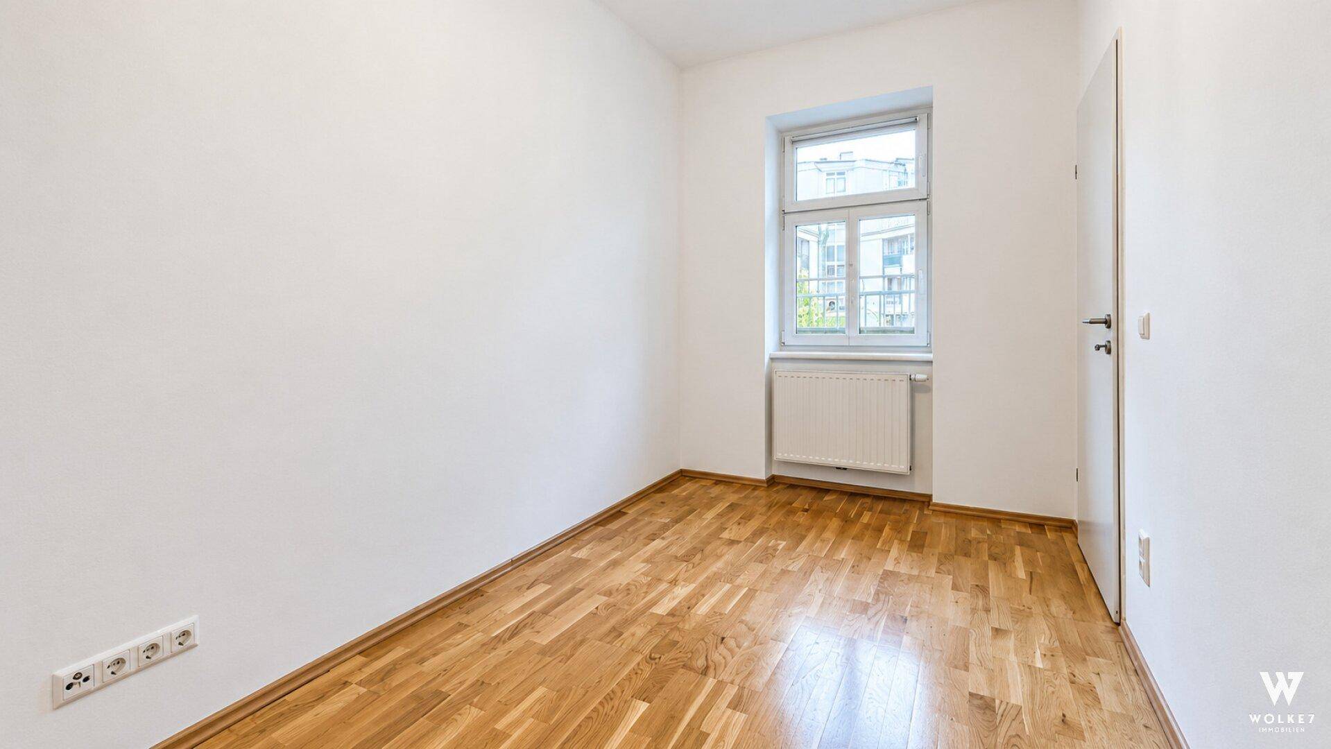 Helles Zimmer mit Parkettboden, einem Fenster und weiß gestrichenen Wänden.