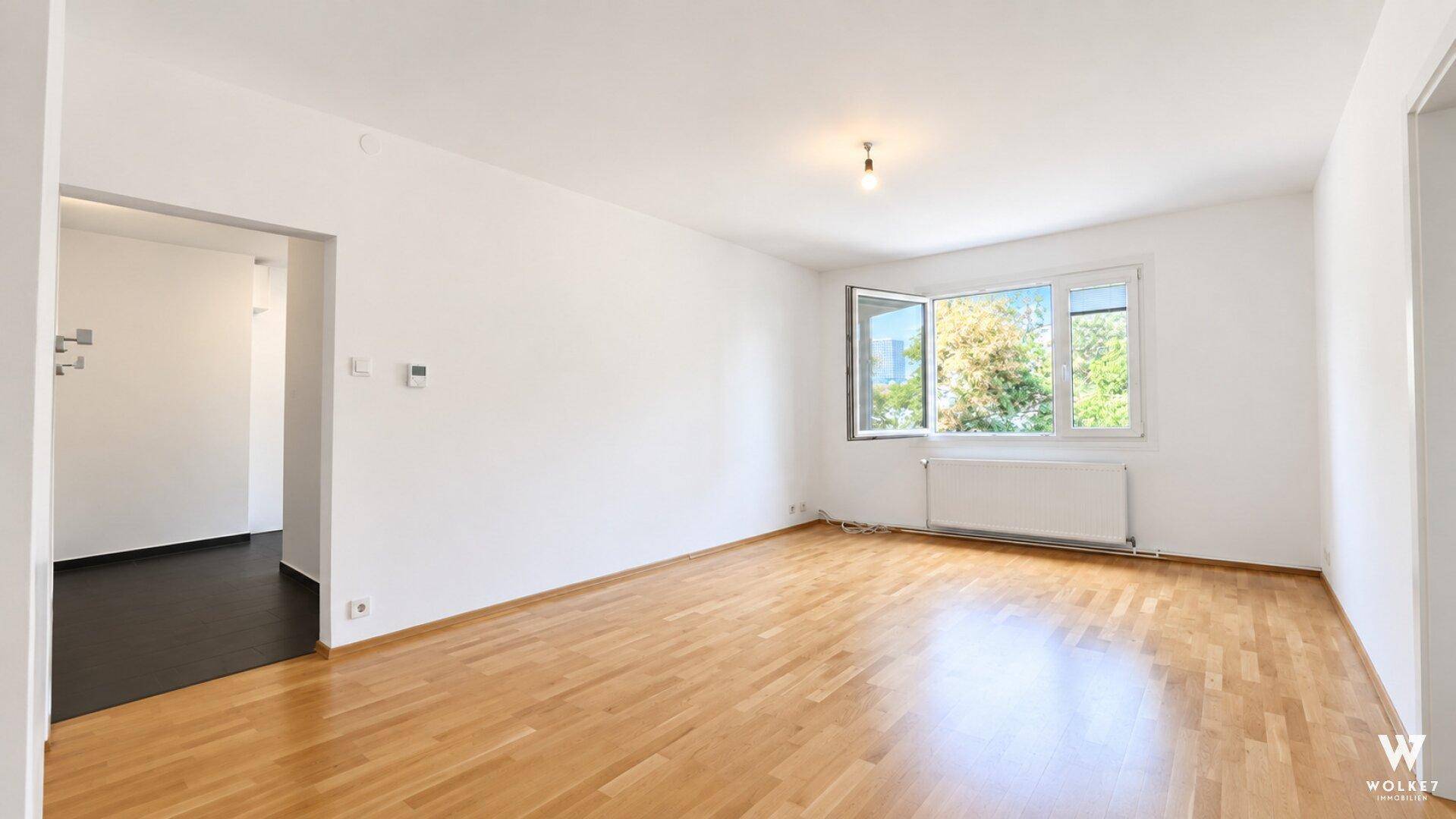Leeres Zimmer mit hellem Parkettboden und Fenster mit Blick ins Grüne.